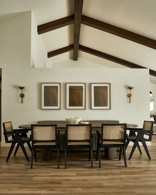 A refined dining room with a long dark dining table surrounded by black-framed cane-back chairs, a built-in upholstered banquette, framed artwork, and sculptural wall sconces. The room has warm timber flooring, cream walls, and a high pitched ceiling with exposed dark timber beams.
