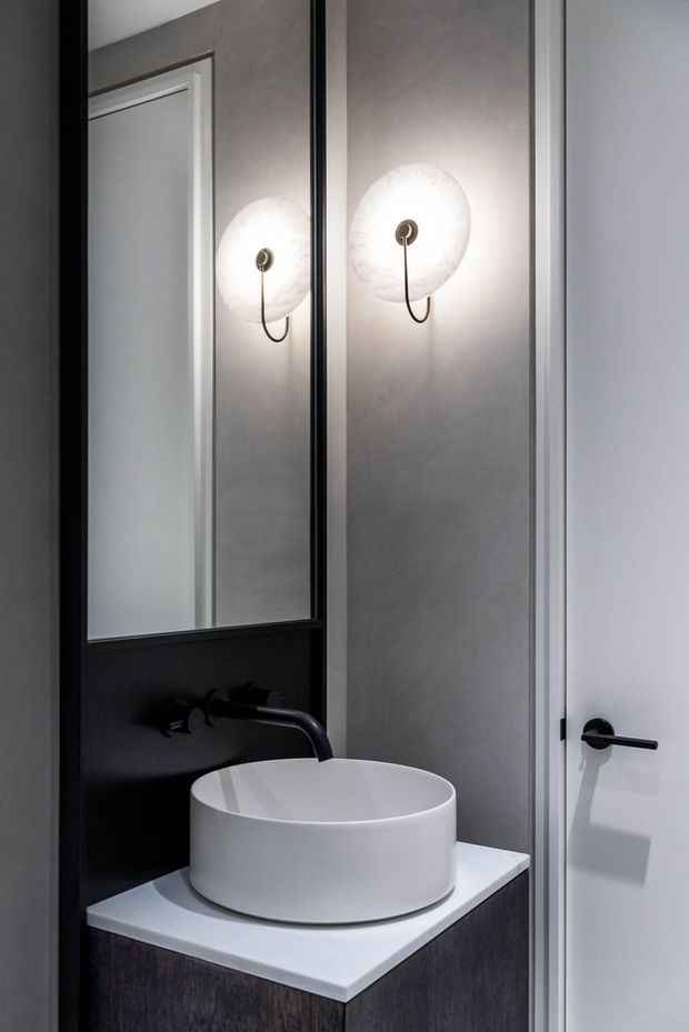 A compact, refined bathroom vanity viewed straight on, featuring a white vessel basin on a white benchtop, dark wood-toned or charcoal vanity cabinetry, a black wall-mounted tap, black-framed mirror, and a sculptural glowing wall sconce. The space has light grey walls, white trim and door, and strong black-and-white contrast.