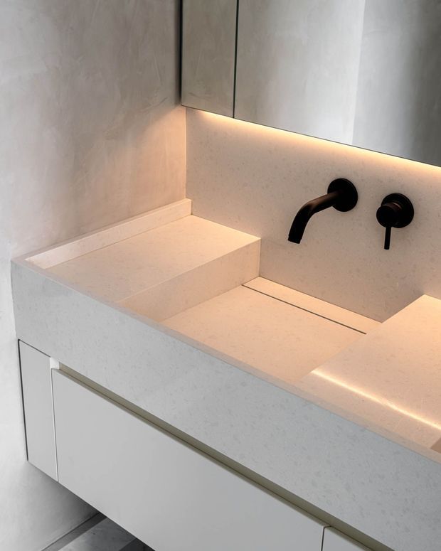 Close-up of a minimalist bathroom vanity with an integrated white stone basin and raised rear ledge. The vanity features pale slab cabinetry, a matching stone splashback and benchtop, a wall-mounted matte black tap and mixer, and warm concealed LED lighting beneath the mirrored wall cabinet. The composition is clean, refined, and contemporary.