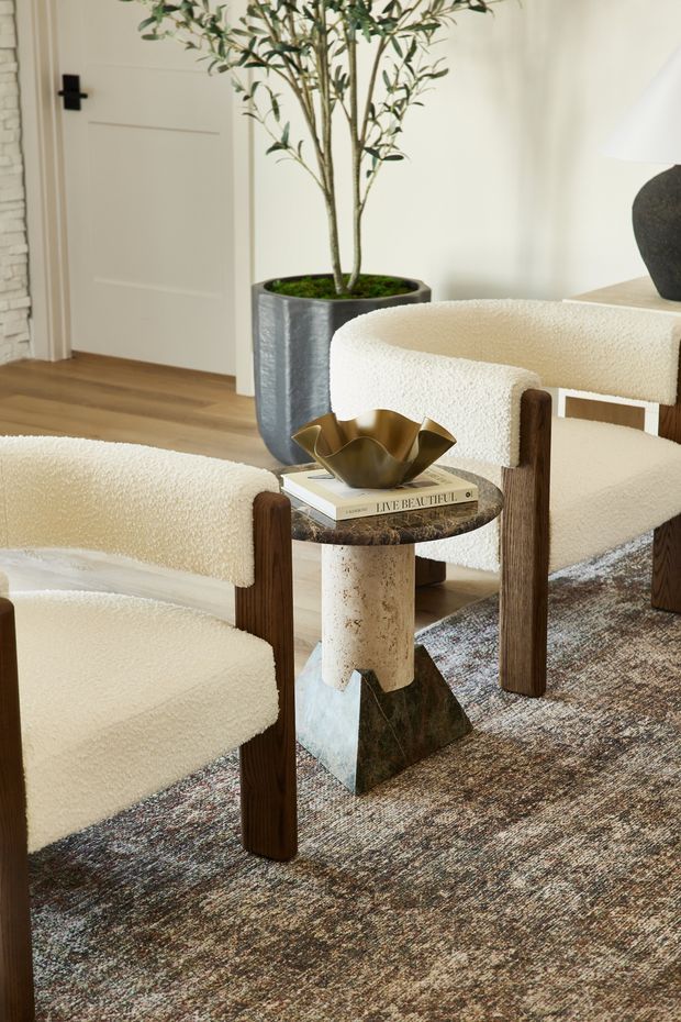 A refined living or sitting area featuring two cream bouclé upholstered lounge chairs with dark timber frames, arranged around a small sculptural side table. The table combines a rounded dark stone top, a textured cylindrical light stone pedestal, and a dark geometric metal base, with a brass decorative bowl and book on top. A large patterned rug, pale timber flooring, white walls, a paneled door, exposed light brickwork, and a tall indoor plant complete the warm neutral interior.
