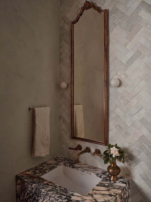 A moody, elegant bathroom vanity featuring a rectangular under-mounted sink set into a dramatic veined natural stone countertop. The wall behind is clad in light herringbone tiles, with a tall ornate dark timber-framed mirror, aged bronze wall-mounted tapware, spherical wall lights, and a towel rail with a cream towel. A decorative metallic vase with white flowers adds a refined accent.