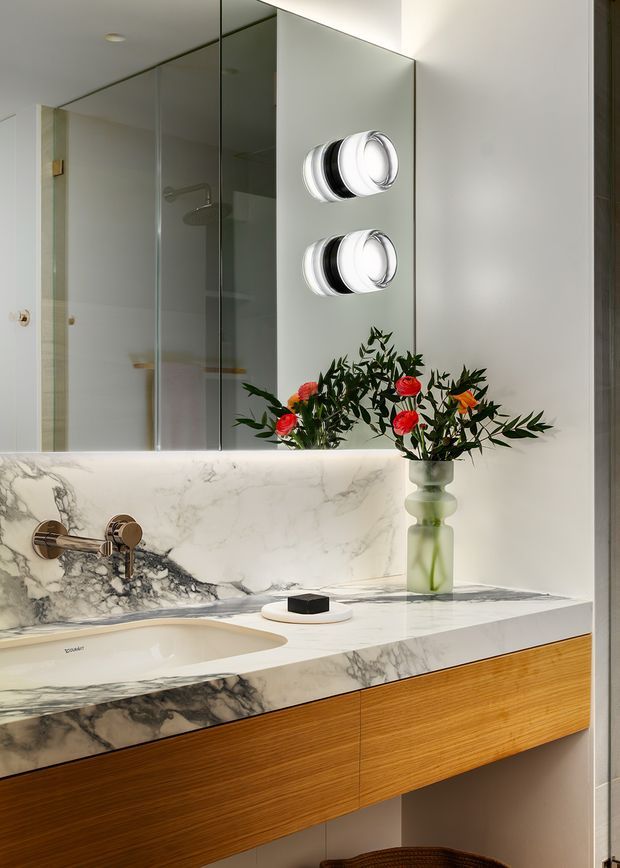 A refined bathroom vanity with a wall-mounted basin set into a dramatic veined marble countertop and backsplash. The floating timber-grain vanity provides warm contrast beneath the pale stone, while a large mirrored wall reflects a glass-enclosed shower. Sculptural chrome wall lights and a green glass vase with red and orange flowers add decorative detail.