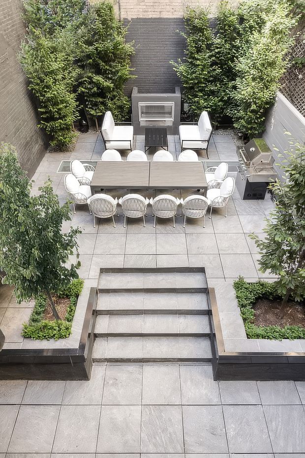 A high-angle view of a formal landscaped courtyard terrace with large grey paving slabs, broad steps, raised planting beds, clipped greenery, and a dining and lounge arrangement. A long outdoor dining table with woven chairs sits centrally, with a compact outdoor kitchen or grill to the right and a built-in outdoor fireplace or heater against the dark textured rear wall. The space has a restrained contemporary design with a neutral grey and white palette and dense green planting.