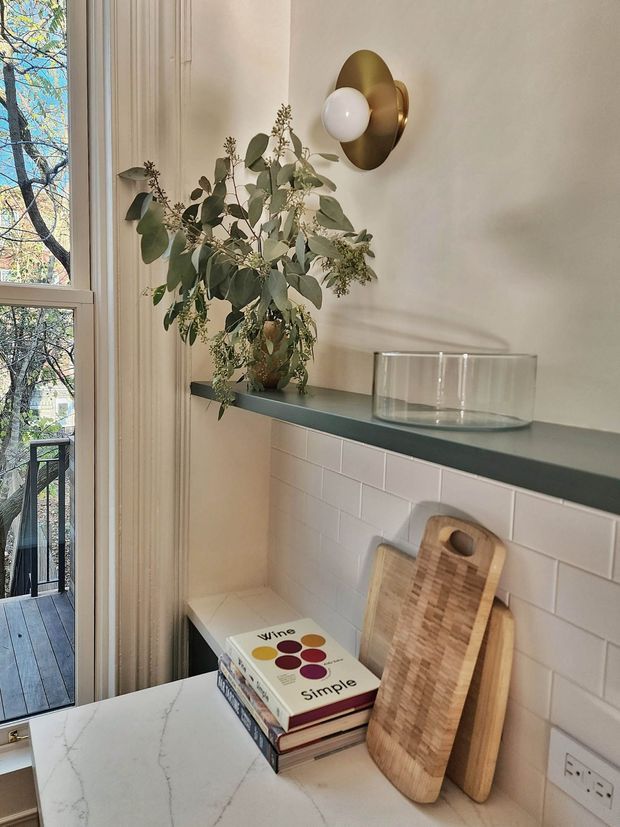 A warmly styled interior vignette beside a tall glazed door or window, featuring a narrow dark countertop or shelf above a white rectangular tiled wall. A brass-and-white globe wall sconce, leafy branch arrangement in a vase, clear glass vessel, stacked books, and wooden cutting boards create a refined, contemporary kitchen or entertaining nook. Natural light and views to a balcony or exterior walkway are visible through the glazing.