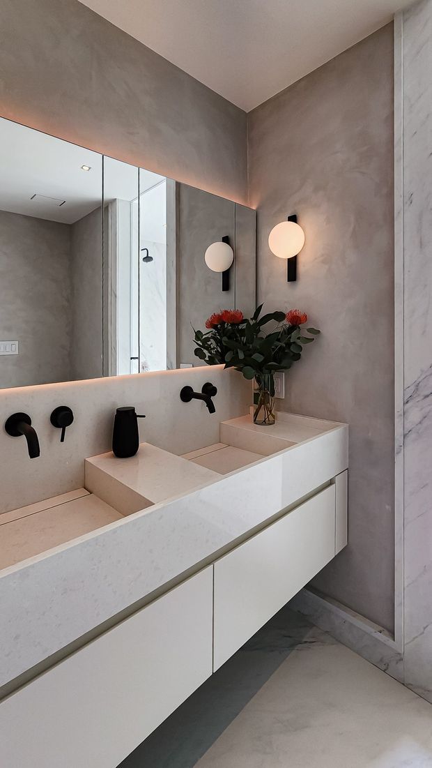 A refined contemporary bathroom featuring a long floating double vanity with pale stone surfaces, integrated basins, matte black wall-mounted tapware, and extensive mirror panels. Warm globe sconces, grey textured plaster walls, and a veined stone wall panel create a calm, luxurious atmosphere.