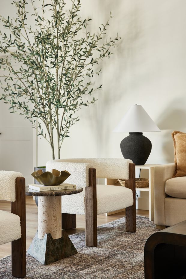 A warmly styled living room with cream boucle lounge chairs, a beige sofa, dark timber chair legs, a sculptural stone-and-metal side table, and a black ceramic table lamp with a white shade. A tall indoor tree adds an organic element, while a textured neutral rug and soft off-white walls create a refined, earthy atmosphere.