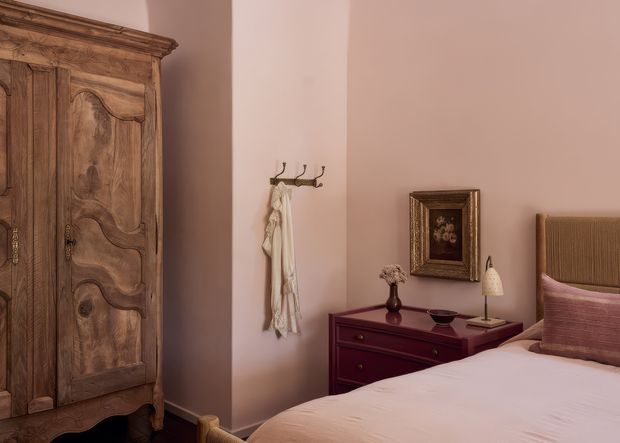 A warm, softly lit bedroom featuring a large ornate rustic wooden armoire, blush-pink walls, a dark burgundy bedside chest, framed artwork, and a neatly made bed with a textured upholstered headboard and pink accent cushion. The room has an elegant traditional character with muted, refined tones.