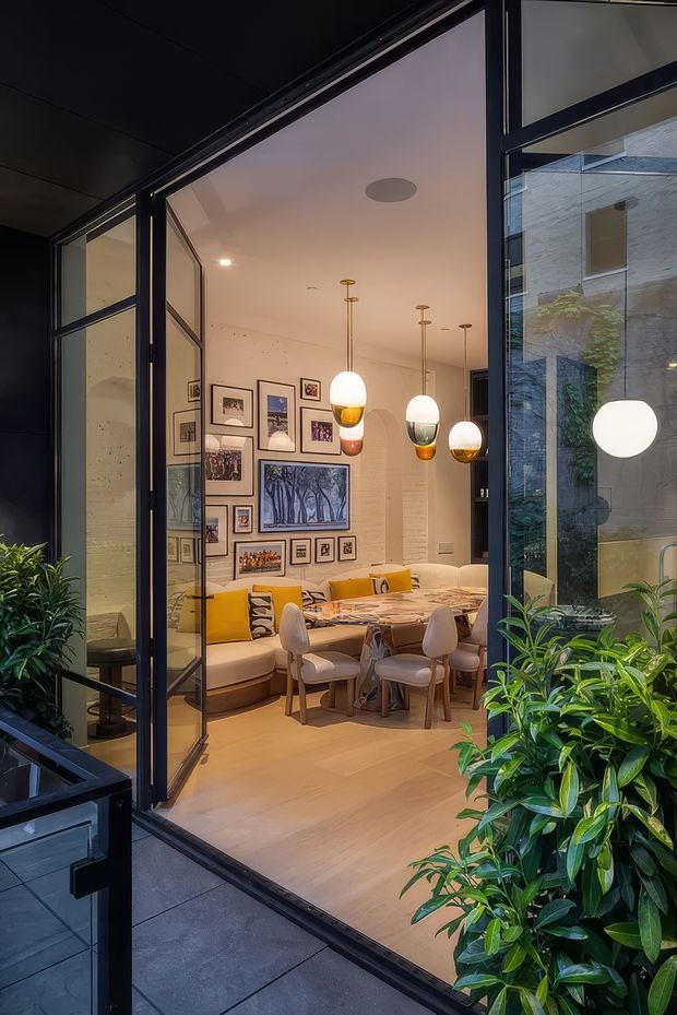 A view from an exterior courtyard or terrace through large dark-framed glazed doors into a warm, highly styled dining and living area. The interior features a timber-toned floor, a dining table with upholstered chairs, a curved built-in banquette with mustard cushions, a gallery wall of framed artwork, and several suspended globe pendant lights. Dense green planting frames the doorway, while reflections reveal the surrounding glazed architectural enclosure.