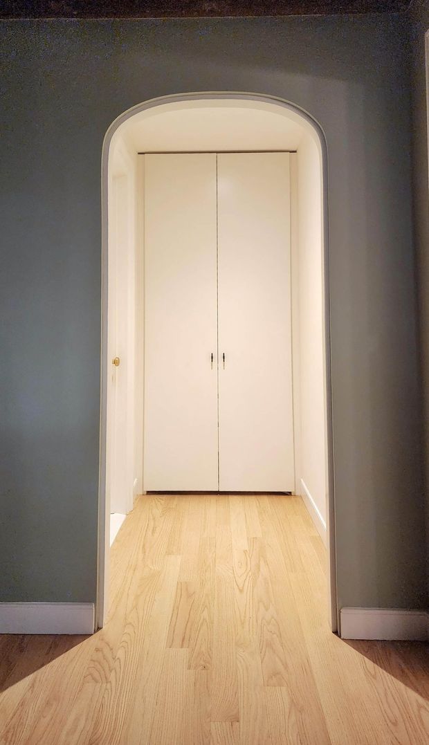 A narrow interior passage framed by a rounded archway, leading to a pair of tall white built-in cupboard doors. The surrounding walls are dark grey with white skirting boards, while the continuous floor is light natural timber. The image has a simple, understated character with warm lighting and strong vertical symmetry.