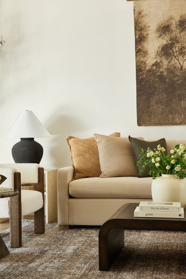 A warmly styled living room with a beige upholstered sofa, layered tan and neutral cushions, a dark sculptural coffee table, textured area rug, and a wood-framed boucle armchair. A black ceramic table lamp, leafy floral arrangement, and large sepia-toned botanical wall artwork add contrast and organic character. The palette is earthy, neutral, and softly refined.
