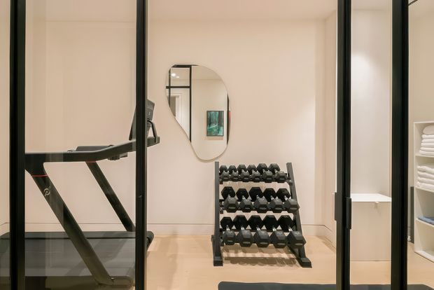A compact home gym viewed through black-framed glass doors, featuring a treadmill in the foreground, a multi-tier dumbbell rack, pale walls, light timber flooring, and built-in open shelving with neatly folded white towels. A softly rounded wall mirror adds visual interest and reflects part of the room.
