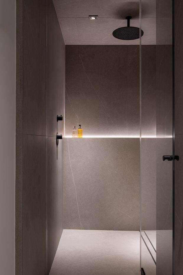 A compact, contemporary walk-in shower enclosed by clear glass, with large warm grey stone-look wall and floor tiles, matte black shower fittings, recessed ceiling lighting, and a thin illuminated shelf niche holding toiletries. The design is minimalist and moody, with clean lines and concealed detailing.