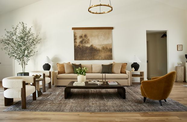 A refined contemporary living room with a cream upholstered sofa, two rounded armchairs, a dark low coffee table, neutral textured rug, timber flooring, and warm earth-toned cushions. A large sepia-toned artwork, sculptural pendant light, table lamps, indoor tree, and restrained decorative objects create a calm, sophisticated setting.