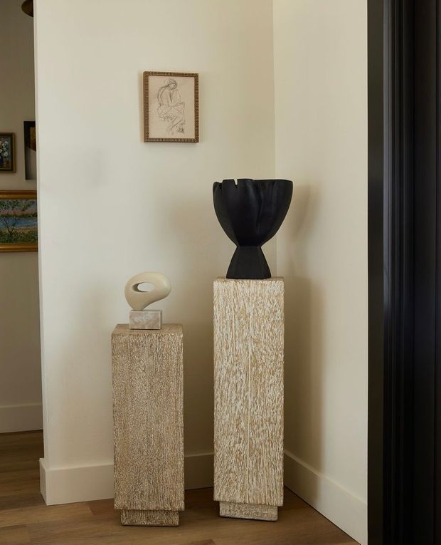 A warmly lit interior vignette in a neutral-toned hallway or entry area, featuring two tall textured pedestal plinths displaying sculptural objects. The cream walls, timber flooring, framed artwork, and dark vertical door or trim create a refined, minimalist composition.