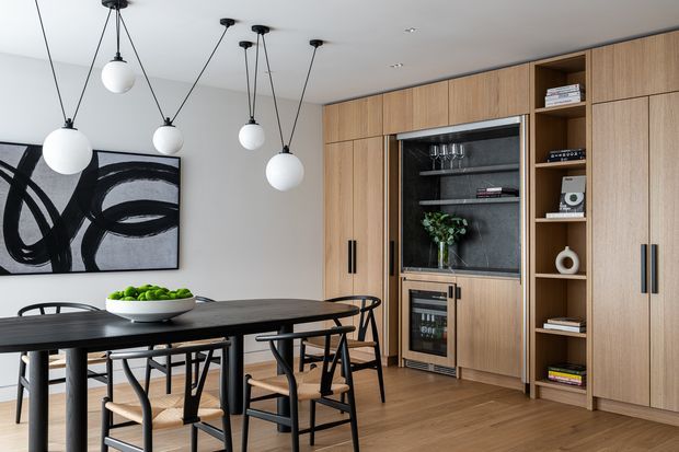 A refined open-plan dining and entertaining space with a dark timber dining table and sculptural black-and-timber chairs beneath multiple spherical pendant lights. A large abstract black-and-white artwork hangs on the pale wall, while a full-height timber built-in wall provides concealed storage, open shelving, a wine refrigerator and display niches. The room has warm timber flooring and a restrained neutral palette.