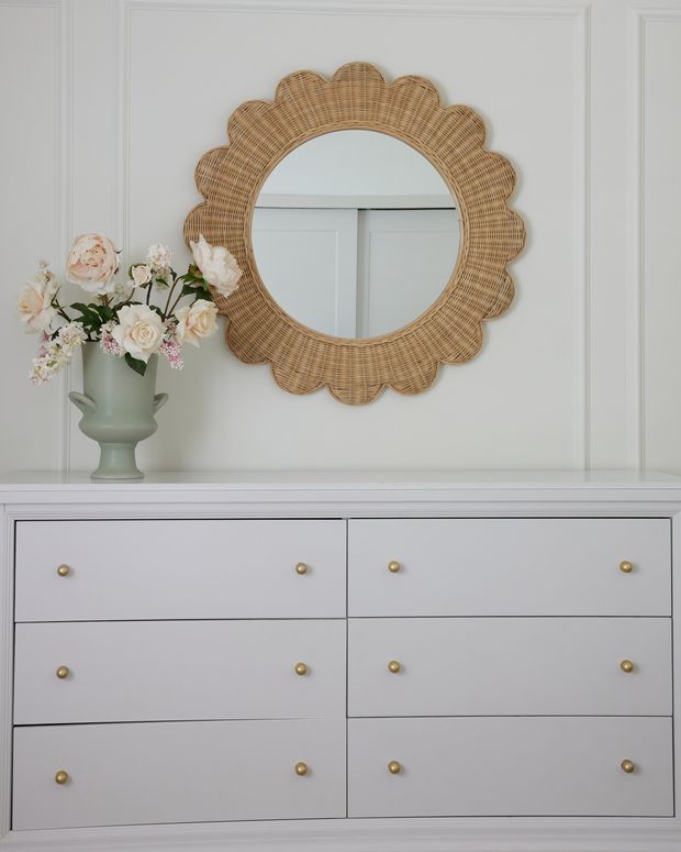 A styled white paneled wall with a large round scalloped woven rattan mirror above a white six-drawer dresser. A pale sage-green vase filled with soft pink and white flowers sits on the dresser, creating a calm, refined bedroom or entryway vignette. The composition uses traditional wall moulding, soft neutral colours, and natural woven texture.