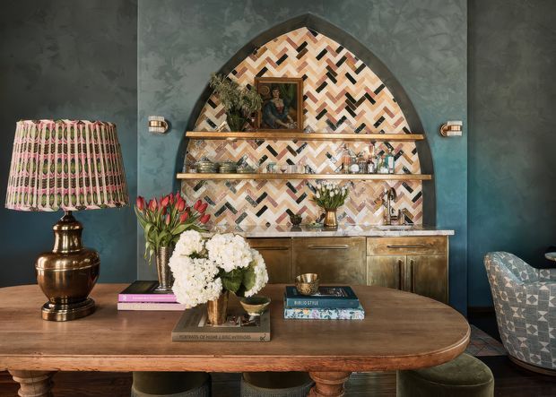 Eclectic, richly layered interior vignette featuring a timber console or dining table, upholstered seating, a brass-toned lamp, decorative objects and flowers. The focal point is an arched recessed alcove lined with a bold geometric herringbone mosaic tile pattern, with open timber shelves and a brass-toned sideboard below. Deep teal textured walls and warm timber, brass and patterned finishes create a dramatic, colourful atmosphere.