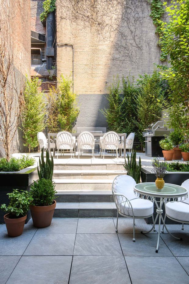 A lush urban courtyard terrace enclosed by tall brick and rendered masonry walls. Broad stone-paved steps lead to an elevated dining area with a white outdoor table and chairs, while a second round bistro table and chairs occupy the foreground. Potted plants, clipped shrubs, climbing vines, and a built-in barbecue create a private garden entertaining space. The image has a refined, traditional urban courtyard character with bright natural light.