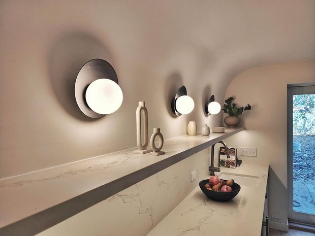 Warm, refined kitchen interior featuring a long pale stone benchtop with subtle veining, a raised splashback or divider, dark undermount sink and gooseneck tap, and minimalist black-and-white wall sconces. Neutral plaster walls, small decorative ceramics, a potted plant, and a glazed exterior door complete the softly lit space.
