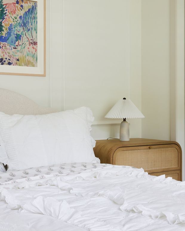 A softly styled bedroom with a white, textured bed and layered bedding, pale green decorative wall panelling, a timber bedside cabinet with woven cane drawer fronts, and a sculptural pleated table lamp. A colourful framed artwork adds a small accent above the bed.