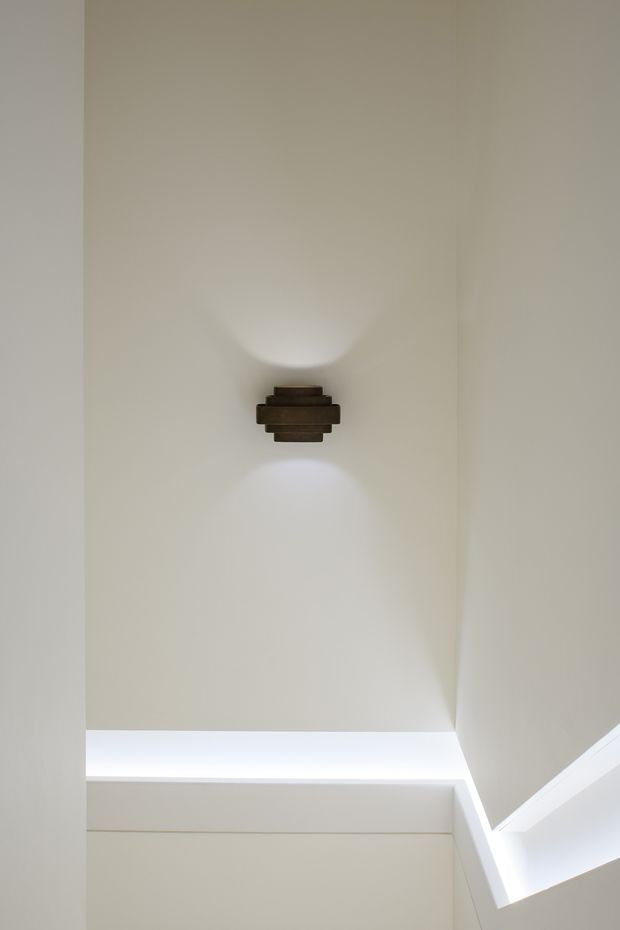 Minimal architectural interior detail showing a warm off-white plastered wall with a centrally mounted dark bronze or brown wall sconce casting soft up-and-down light. A bright linear ceiling or clerestory opening is visible at the lower edge, emphasizing the tall, clean-lined vertical composition.