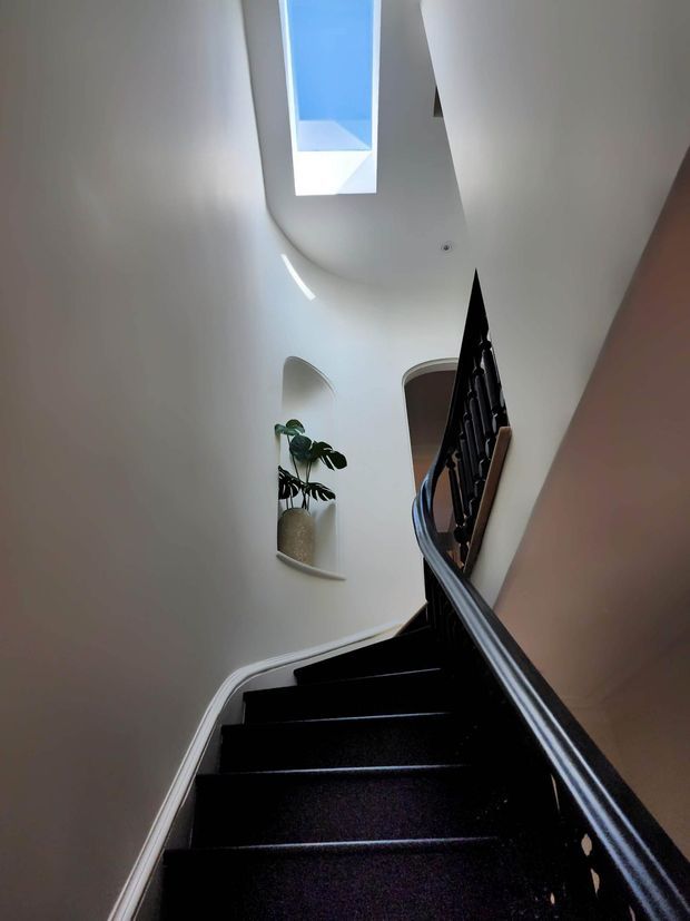 A narrow interior stairway viewed looking upward, with dark carpeted steps and a dark timber or painted handrail and balustrade. The stair curves around a tall white plastered stairwell with arched wall niches, one containing a potted plant, and a skylight bringing in blue daylight from above.