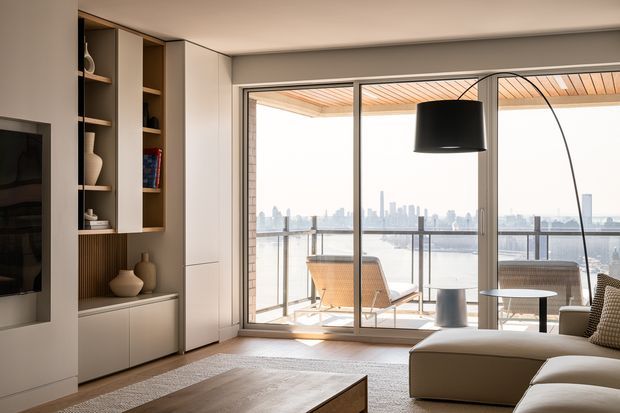 A refined contemporary living room with a built-in media and display wall, pale floor-to-ceiling cabinetry, warm timber backing, a neutral sectional sofa, and a low timber coffee table. Large sliding glass doors open to a balcony with outdoor seating and expansive waterfront city views. A curved black floor lamp provides a strong sculptural accent, while the timber-lined balcony ceiling adds warmth.