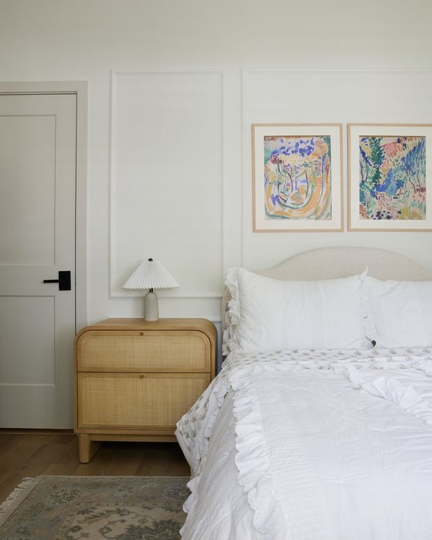 A bright, softly styled bedroom with white decorative wall panelling, a paneled white door with black hardware, and a curved upholstered bed dressed in layered white bedding. A natural cane-front bedside cabinet supports a pleated table lamp, while two colourful framed artworks hang above the bed. Timber flooring and a muted patterned rug add warmth.