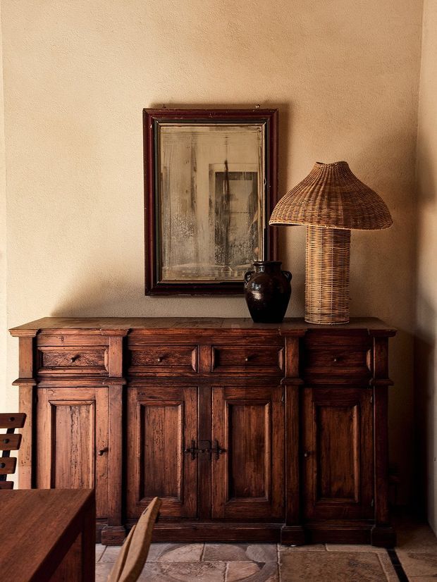 A warmly lit interior vignette featuring a large dark timber paneled sideboard against a textured beige plaster wall. A framed artwork, dark ceramic vase, and woven rattan table lamp sit on top, with partial timber dining furniture and a stone or terracotta-toned tiled floor visible in the foreground. The composition has a rustic, heritage-inspired character with rich wood tones and subdued earthy colours.