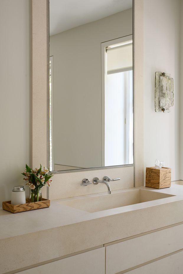 A bright, minimalist bathroom vanity with a large frameless mirror, integrated rectangular basin, wall-mounted chrome faucet, and pale stone or solid-surface benchtop and splashback. The vanity has simple light-coloured drawers, while a small timber tissue box and tray with flowers add warm natural accents. A window with a light roller blind is reflected in the mirror.