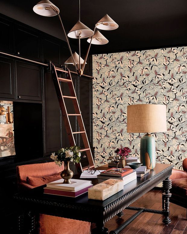 A richly layered, moody living room with black built-in cabinetry, a tall wooden library ladder, bold botanical wallpaper, and a large dark wood coffee table. Coral upholstered seating, sculptural table lamps, books, flowers, and decorative objects create an eclectic, refined interior with strong contrast and vintage character.
