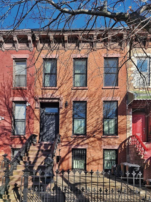 Street-facing view of a narrow attached red-brick townhouse row, featuring ornate Victorian-style cornice detailing, tall dark-trimmed sash windows, decorative wrought-iron security grilles and fencing, and steep front stoops with elaborate iron railings. Leafless tree branches cast shadows across the warm brick facade under a clear blue sky.