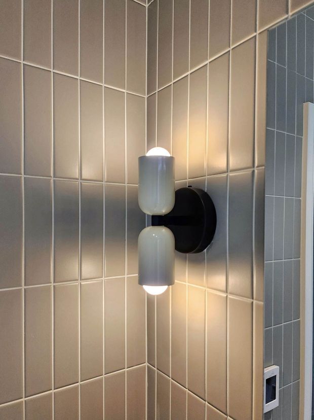 Close-up of a wall-mounted double globe light fixture in a tiled interior corner. The warm light illuminates beige-grey vertically arranged rectangular wall tiles with contrasting grout, while a darker grey tiled wall and a small control panel are visible at the edge.
