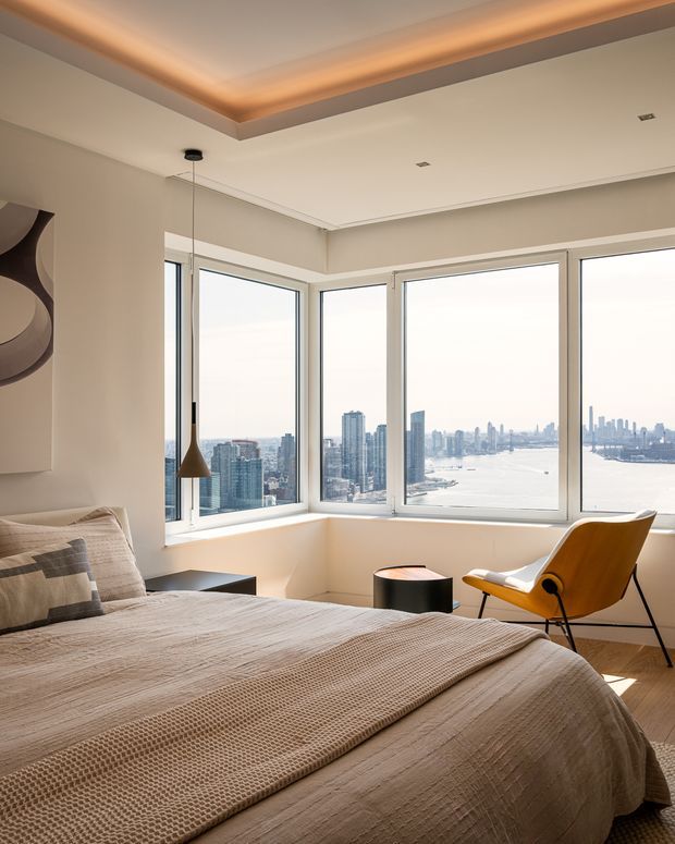 A bright, refined bedroom with expansive corner windows overlooking a dense waterfront city skyline. The room features a large upholstered bed with layered neutral bedding, a sculptural pendant light, a mustard-yellow lounge chair, a small dark side table, pale walls, light timber flooring, and a recessed ceiling detail with warm cove lighting.