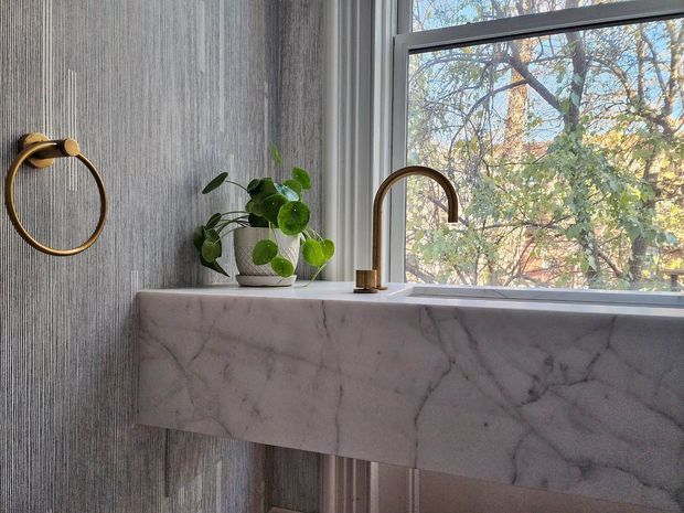 Close-up of a refined bathroom vanity beneath a large window. The floating vanity has a pale veined stone benchtop and fascia, a white vessel basin, and a curved brass tap. Vertically textured grey wall tiles, a brass towel ring, and a leafy potted plant complete the calm, natural-looking composition. Trees are visible outside the window.