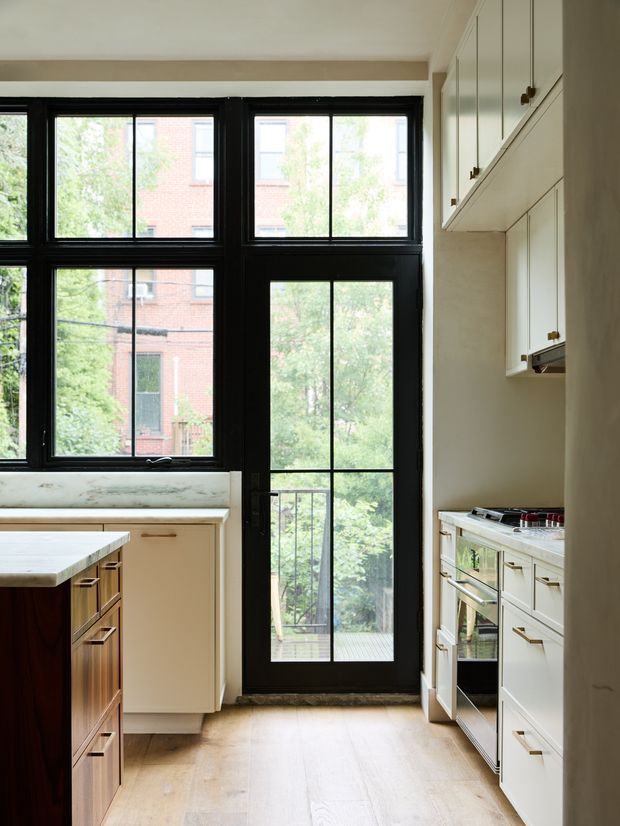 Bright, narrow kitchen with cream-painted cabinetry, a central island, pale timber flooring, and black-framed industrial-style windows and glazed rear door. The room includes a light stone benchtop, integrated cabinetry, and a built-in stainless-steel oven and cooktop. Natural light and leafy urban views create an airy, refined atmosphere.