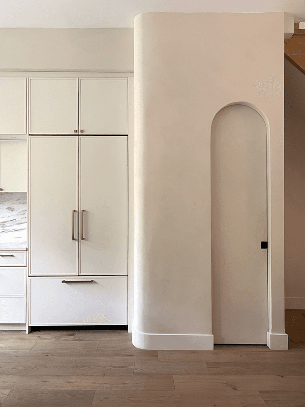 A warm, minimalist interior detail showing a built-in white kitchen cabinet bank with concealed storage, long vertical metal handles, and a pale stone worktop at left. A softly rounded plaster partition wall creates an arched doorway, with light timber flooring and neutral painted surfaces throughout.