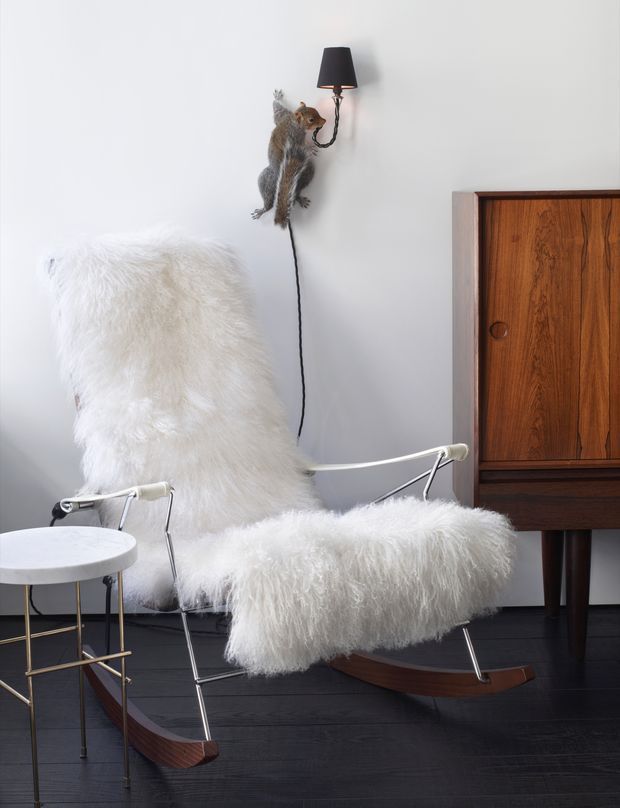 A styled interior vignette featuring a white shaggy sheepskin-covered lounge chair with a chrome tubular frame and curved dark wood rockers. A small round side table sits beside it, while a mid-century wooden cabinet, wall-mounted black lamp, and playful squirrel wall sculpture complete the scene. The room has a pale grey wall and dark timber flooring, creating a high-contrast, curated aesthetic.