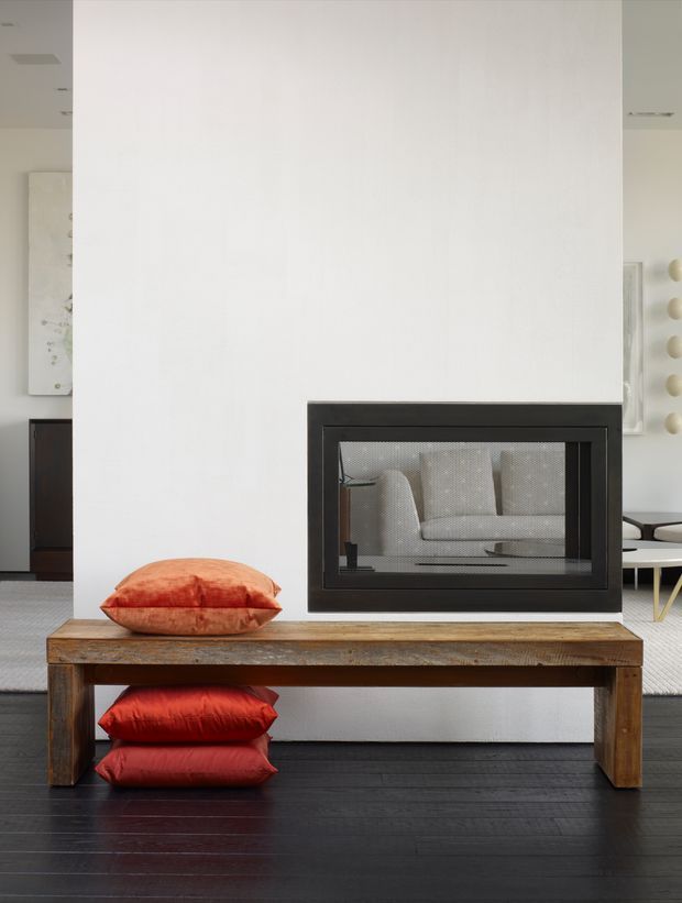 Interior view of a minimalist living area centered on a white textured feature wall with a built-in black-framed fireplace. A rustic timber console bench sits in front, with red-orange cushions, dark timber flooring, and partial views of adjacent furniture and artwork.