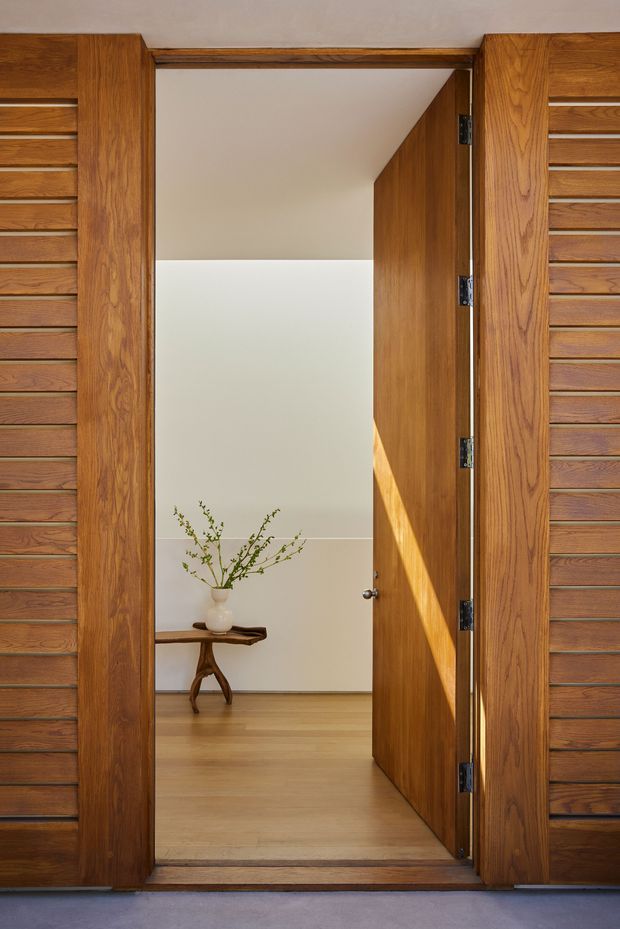 A close-up architectural view of a timber-framed entry doorway with a large open wooden door. The doorway is surrounded by rich warm wood panels with horizontal slatted detailing, opening into a simple bright interior hallway or entry space. Inside, pale timber flooring, white walls, and a rustic live-edge console table with a small vase and branches are visible. Strong sunlight falls across the open door, creating a warm, refined and welcoming composition.
