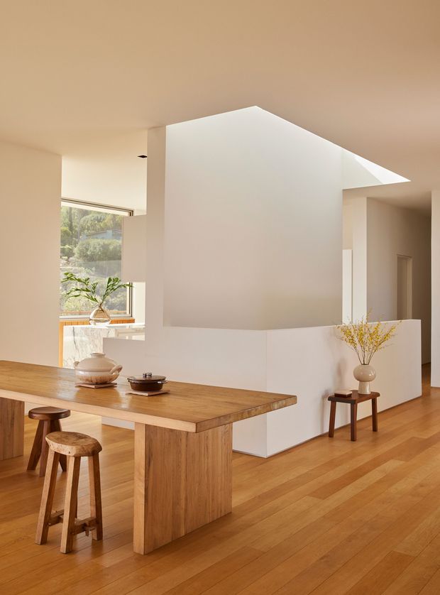 Warm, minimalist open-plan interior with a large solid timber dining table and stools in the foreground. A sculptural white geometric partition or stair volume dominates the centre, creating a strong angular ceiling form. Timber flooring continues throughout, with a small side table, vase, and restrained decorative objects. A large window brings in natural light and views of greenery.