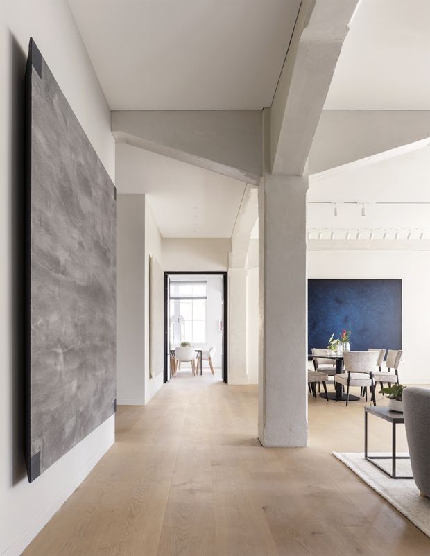 Bright contemporary open-plan interior viewed from a hallway or circulation area, with pale timber flooring, white plaster walls and ceilings, substantial exposed structural columns and beams, and a dining/living area visible to the right. A large dark textured wall artwork or feature panel contrasts with the otherwise restrained palette. The space appears airy and refined with high ceilings and strong architectural sightlines.