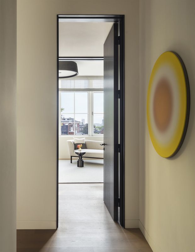 A view down a refined residential hallway toward a bright living room, framed by a dark timber or painted door opened into the room. The hallway has warm beige walls, dark wood flooring, simple white skirting, and a large circular yellow wall artwork. Beyond the doorway is a light-toned lounge with a sofa, small round coffee table, pendant light, expansive glazing, and an urban outlook. The image has a clean, contemporary, gallery-like composition with strong axial framing and a contrast between the darker corridor and bright living area.