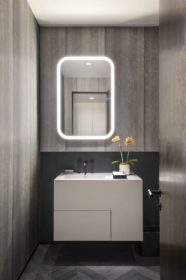 A compact, refined bathroom with a floating light-grey vanity, integrated white basin, matte black tapware and backsplash, and a rounded rectangular backlit mirror. Dark vertically panelled walls and a dark herringbone tile floor create a dramatic, contemporary interior. A small orchid adds a natural accent, while a partially open door and reflected doorway are visible.