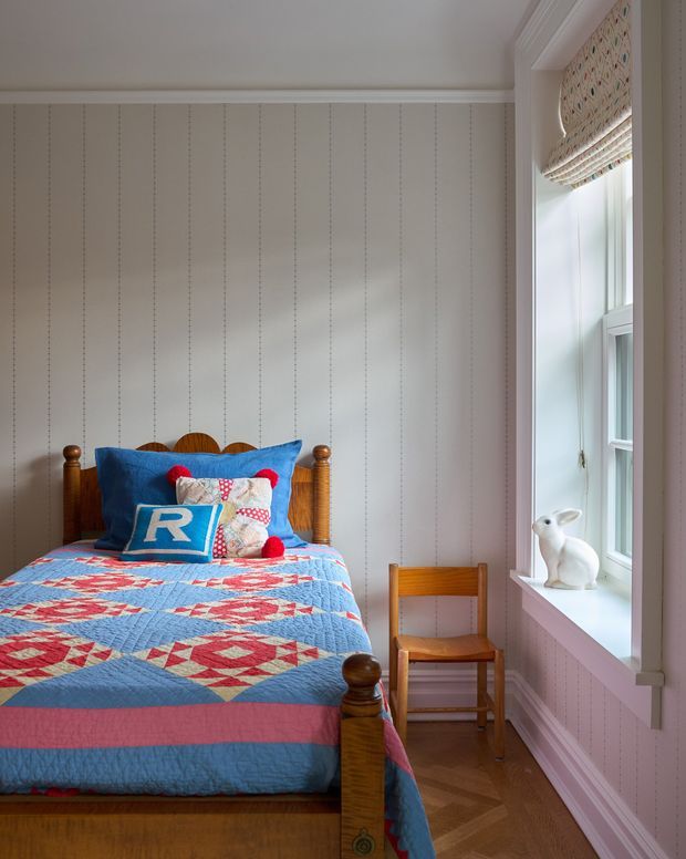 A bright, compact traditional bedroom with a wooden bed, colourful blue-and-red patchwork quilt, patterned pillows, and a small wooden chair. The room has white vertical wall panelling with dotted seams, white trim, timber parquet flooring, and a tall window with a patterned Roman blind. A small white rabbit ornament sits on the windowsill.