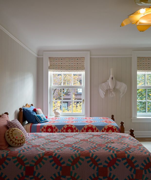 A cozy bedroom with two single beds dressed in colourful patterned quilts. The room features pale vertical wall panelling, white decorative window trims, patterned roller blinds, timber bed frames, and a sculptural white wall-mounted decoration between the windows. Warm sunlight enters through the multi-pane windows, while a playful pendant light is partially visible overhead.