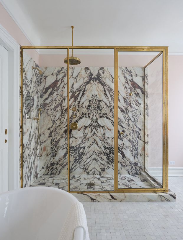 Luxurious bathroom featuring a large walk-in glass shower enclosed in a brass-framed partition. The shower walls and floor use dramatic bookmatched veined marble, complemented by pale pink walls, white decorative moulding, and a freestanding white bathtub in the foreground. The overall design is highly ornate and refined.