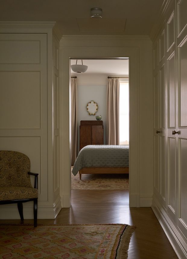 A refined residential hallway or landing viewed toward an open bedroom doorway. The corridor features extensive cream painted wall panelling and moulded trim, polished timber flooring, built-in paneled cabinetry or wardrobes along the right wall, and a patterned area rug. A patterned upholstered occasional chair sits to the left. Through the doorway, a calm bedroom is visible with a timber bed, pale quilt, curtains, timber cabinet, and decorative mirror.