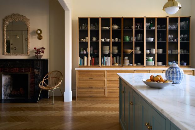 A refined open-plan kitchen and dining display area with a large marble-topped island, extensive dark timber open shelving and drawer cabinetry, and a decorative fireplace visible in the adjoining space. The room features herringbone timber flooring, warm neutral walls, brass lighting and hardware, curated tableware, and a rattan accent chair. The overall look blends traditional architectural detailing with modern classic interior styling.