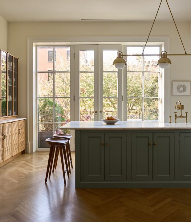 Bright, refined kitchen with a large dark green island, white stone benchtop, brass bridge faucet, timber bar stools, and full-height French doors opening to a leafy exterior. The room features warm herringbone timber flooring, pale walls, glazed cabinetry, and a simple suspended linear pendant with globe shades.